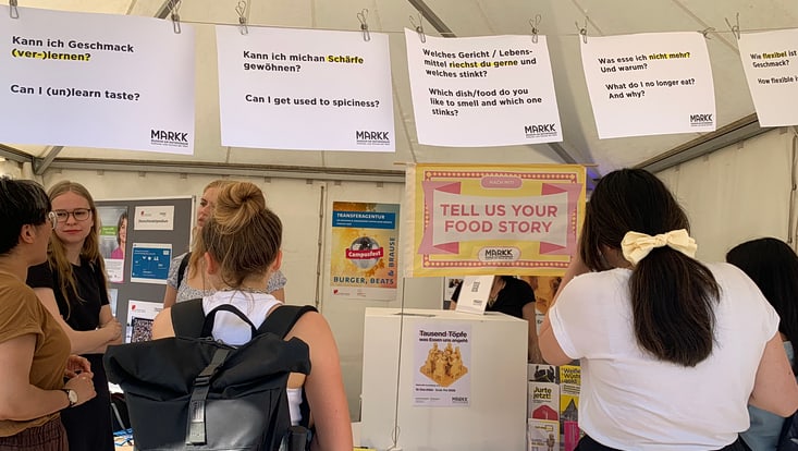 02-05-vernetzung-festivals-ebene-2-slider-campusfest-cucaser-21-733x414 Blätter mit verschiedenen Aufschriften angeheftet an einer Wäscheleine: „Kann ich Geschmack (ver-)lernen? Kann ich mich an Schärfe gewöhnen?“ Und andere. Davor Personen, die sich unterhalten und den Stand betrachten