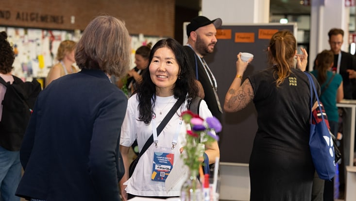2025-09-20-cmc-t-03-alena-zielinski-(81)-733x414 Zwei Personen unterhalten sich, im Hintergrund sind weitere Menschen im Gespräch