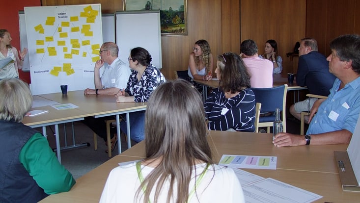 02-06-vernetzung-dialogforen-slider8-2023.06.08-df2-norderstedt-25-733x414 Teilnehmende des Dialogforum Norderstedt sitzen an verschiedenen Tischen und hören der Moderatorin zu, die etwas an einer Pinnwand mit verschiedenen Moderationskarten erklärt