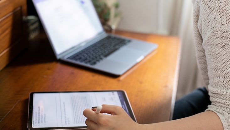 210125-online-studieren-7390-733x414 Person sitzt an einem Holztisch und bedient ein Tablet mit einem Stift, im Hintergrund steht ein aufgeklappter Laptop