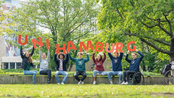 uni-hh-10-2021-1281-733x414 Sieben junge Menschen sitzen auf einer Mauer im Park und halten rote Buchstaben hoch, die zusammen "UNI HAMBURG" ergeben