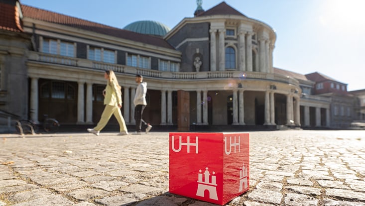 Ein roter UHH-Würfel liegt in der Sonne vor dem Hauptgebäude. Im Hintergrund gehen zwei Menschen.