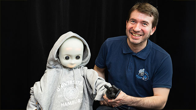 Mann mit kurzen braunen Haaren sitzt neben einem Roboter