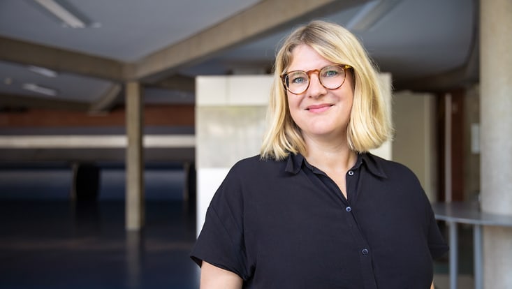 anja-riesel Frau mit hellblonden, schulterlangen Haaren und Brille lächelt in die Kamera. Sie trägt eine schwarze Bluse mit kurzen Ärmeln