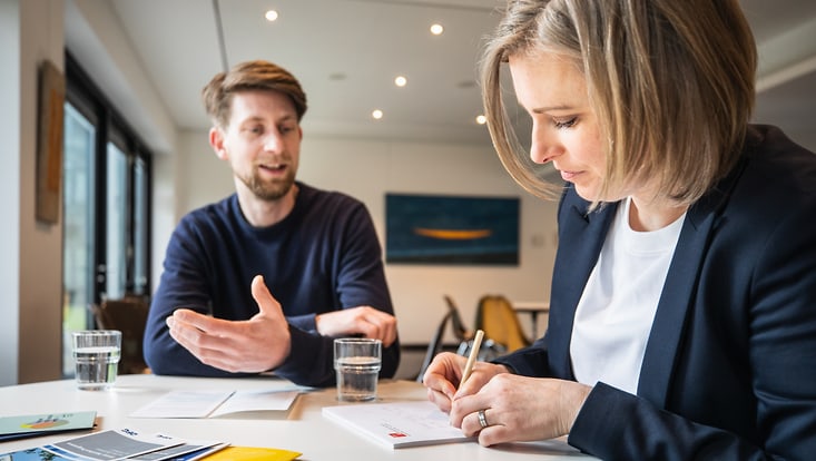 Zwei Personen unterhalten sich an einem Schreibtisch. Eine Person redet und die andere Person schreibt.