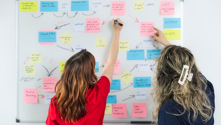 Two women stand in front of a whiteboard, organizing colorful cards.
