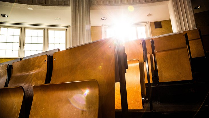 leere Stuhlreihen in einem Hörsaal, durch ein Fenster scheint ein Sonnenstrahl