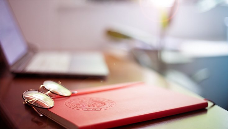 Schreibtisch, auf dem ein rotes Notizbuch der Uni Hamburg liegt. Darauf eine Brille und daneben ein Laptop.