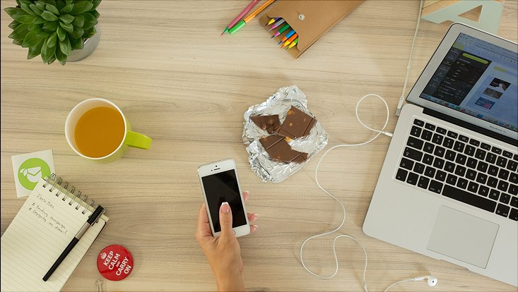 Man sieht eine Hand mit Smartphone, einen Laptop, einen Becher, eine offene Tafel Schokolade