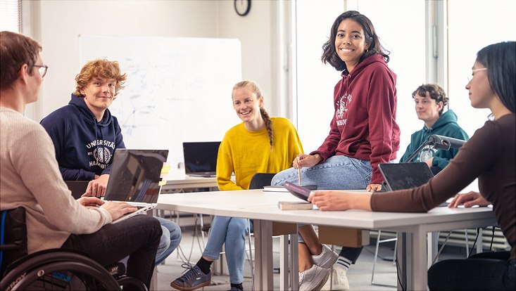 Studierende sitzen um einen Tisch. Auf dem Tisch sitzt eine Studentin