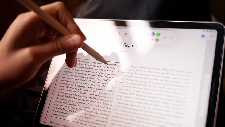 Eine Hand mit Stift. Im Hintergrund ein Tablet mit Text