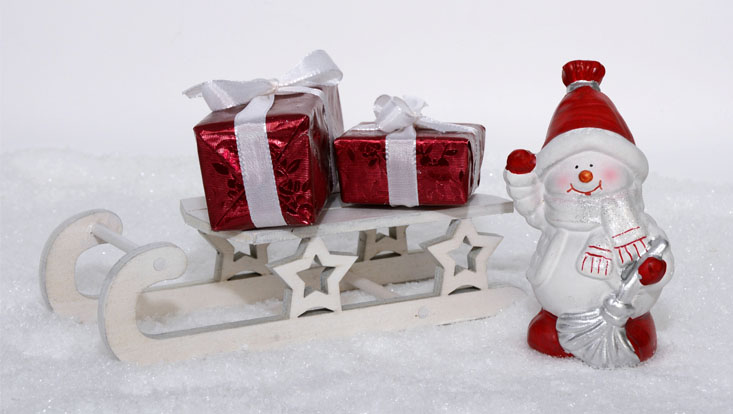 Das Bild zeigt ein Modell von zwei roten Geschenke auf einem Holzschlitten, daneben steht eine Weihnachtsmann-Figur. Der Hintergrund ist weiß und der Boden aus Schnee.