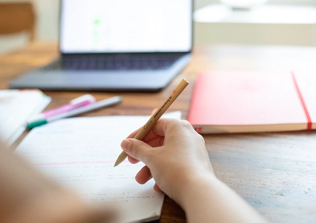 Eine Person schreibt etwas in ein Notizbuch, im Hintergrund ein Laptop