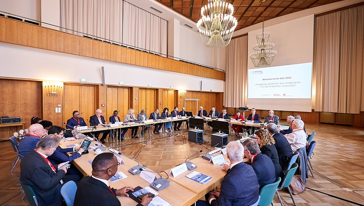 GUC 2023 Menschen sitzen in einem Saal an Tischen, die in einem Viereck angeordnet sind. Im Hintergrund ist eine Power-Point-Präsentation abgebildet.