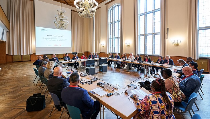 GUC 2023 Menschen sitzen in einem Saal an Tischen, die in einem Viereck angeordnet sind. Im Hintergrund ist eine Power-Point-Präsentation abgebildet.
