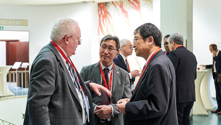 GUC 2023 Drei Personen in Anzügen sprechen miteinander. Im Hintergrund befinden sich weitere Personen.