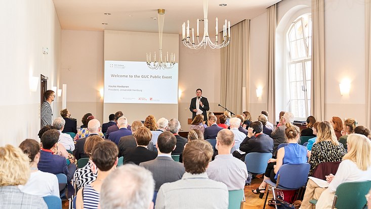GUC 2023 Universitätspräsident Hauke Heekeren spricht auf einer Bühne. Hinter ihm ist eine Power-Point-Präsentation abgebildet und vor der Bühne sitzt das Publikum.