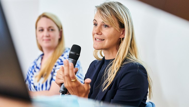 GUC 2023 Eine Frau spricht und hält ein Mikrofon in der Hand. Im Hintergrund sitzt eine weitere Frau.
