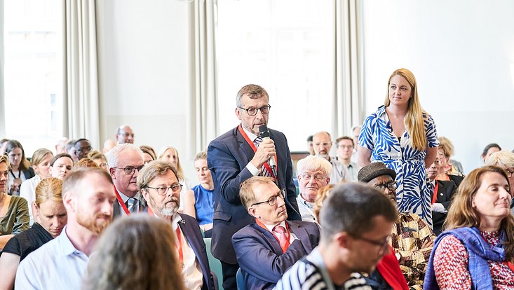 GUC 2023 Zwei Personen stehen zwischen einem sitzenden Publikum. Der Mann spricht und hält ein Mikrofon in der Hand.