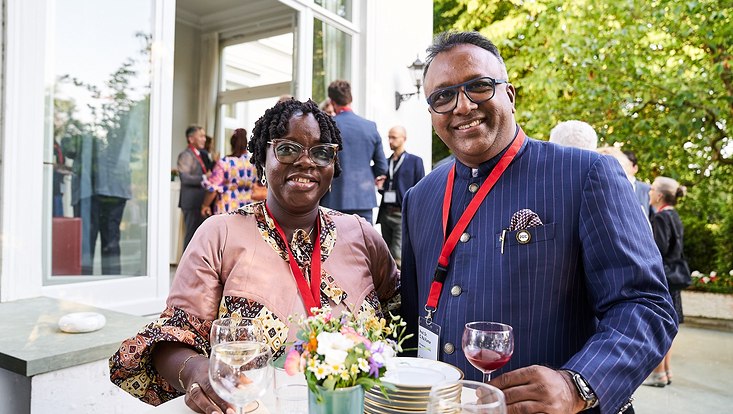 GUC 2023 Eine Frau und ein Mann stehen an einem Tisch. Auf dem Tisch stehen Teller und Gläser. Im Hintergrund stehen weitere Personen.