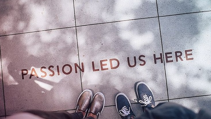 Zwei Paar Füße von Oben Fotografiert auf Betonboden. In der Mitte der Schriftzug auf Englisch "Passion led us here"