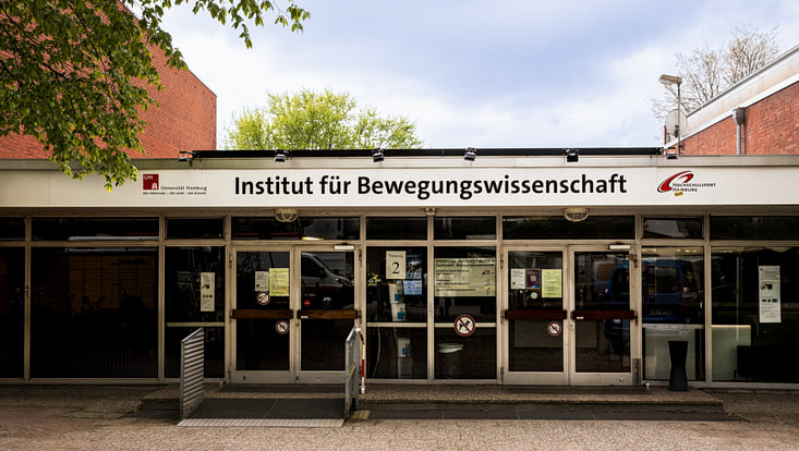 Auf dem Bild sieht man den Eingang zu einem Gebäude. Oben steht: „Institut für Bewegungswissenschaft“. Das gehört zur Universität Hamburg. Der Eingang hat Glas­türen. Es gibt eine Rampe für Rollstuhlfahrer. . Das Gebäude sieht modern und offen aus.