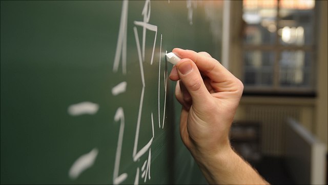 Eine Hand schreibt mit Kreide eine Formel an eine Tafel.