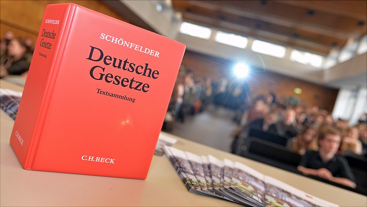 Im Vordergrund ist ein rotes Gesetzbuch "Schönfelder" zu sehen, im Hintergrund sitzen Studierende im Hörsaal Recht.