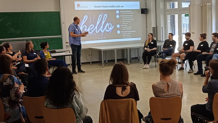 Die Workshopteilnehmenden sitzen im Kreis, der Moderator eröffnet die Veranstaltung. Auf der Präsentation an der Wand steht "hello"