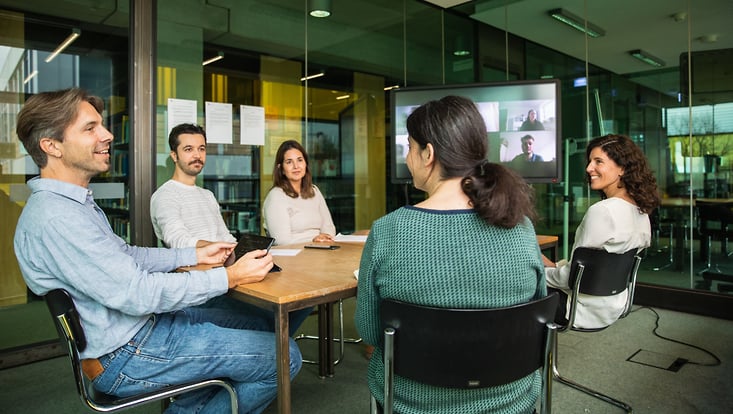 Fünf Menschen unterhalten sich an einem Konferenztisch. Im Hintergrund ist außerdem ein Zoommeeting zugeschaltet.