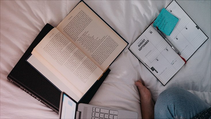 Man sieht Person von oben, die an einem Laptop arbeitet und ein aufgeschlagenes Buch neben sich liegen hat