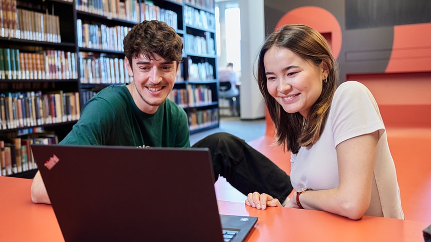 Ein Student und eine Studentin sitzen in einem Lernraum mit Bücherwand vor einem Laptop.