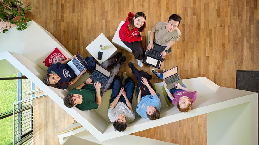 In der Vogelperspektive zeigt das Bild eine Sitzecke mit sieben Studierenden, die Laptops auf den Knien haben und nach oben in die Kamera schauen