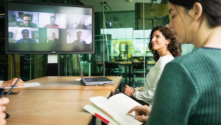 Man blickt in einem Raum in eine hybride Veranstaltung: rechts sitzen PErsonen an einem Tisch, links sieht man einen großen MOnitor auf dem weitere Personen digital zugeschaltet sind.
