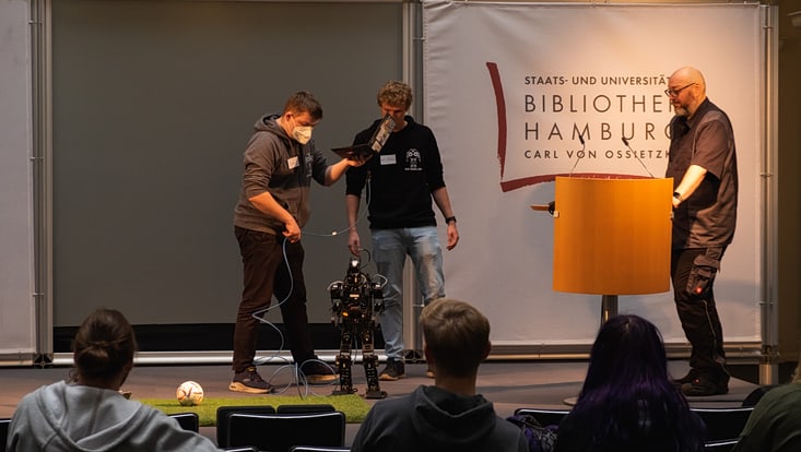 zwei Studierende präsentieren einen laufenden Roboter auf einer Bühne. Daneben steht ein Moderator an einem Rednerpult.