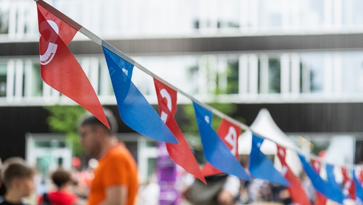 blaue und rote Wimpelchen wehen an einer Leine über den Campus. Im Hintergund sind unscharf Menschen zu sehen.