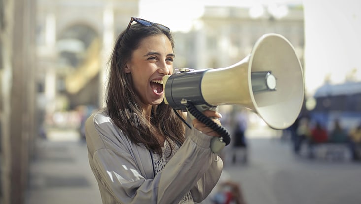A young woman talks loudly into a megaphone, streets and buidlings are blurred but visible behind her