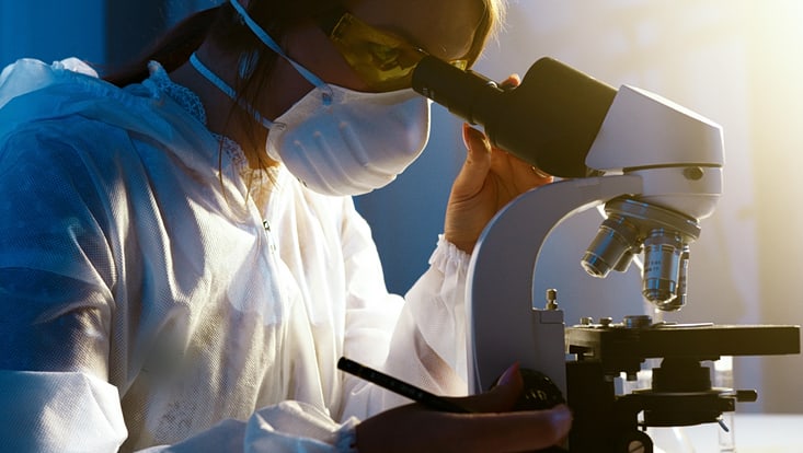A young woman looks into a microscope, wearing laboratory clothing and a face mask
