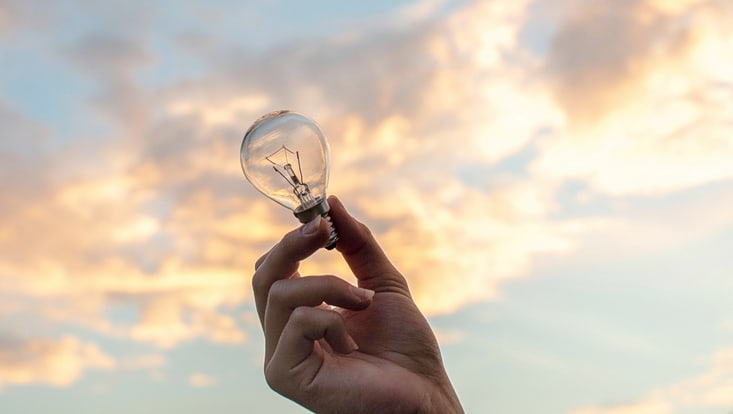 A hand holding a light bulb, in the background reddish-orange evening sky.