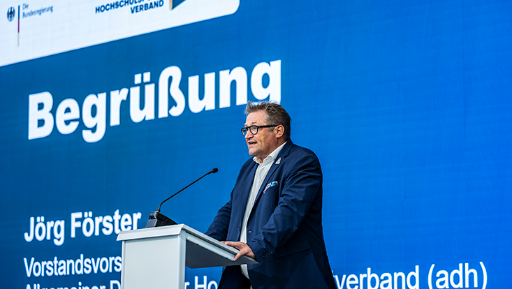 Mann spricht an Redepult vor blauer Leinwand