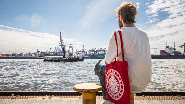 Ein Student sitzt an einem sonnigen Tag an den Landungsbrücken in Hamburg. Er schaut auf den Hafen und die großen Kräne. Er trägt einen roten Leinenbeutel mit dem Aufdruck der Universität Hamburg.
