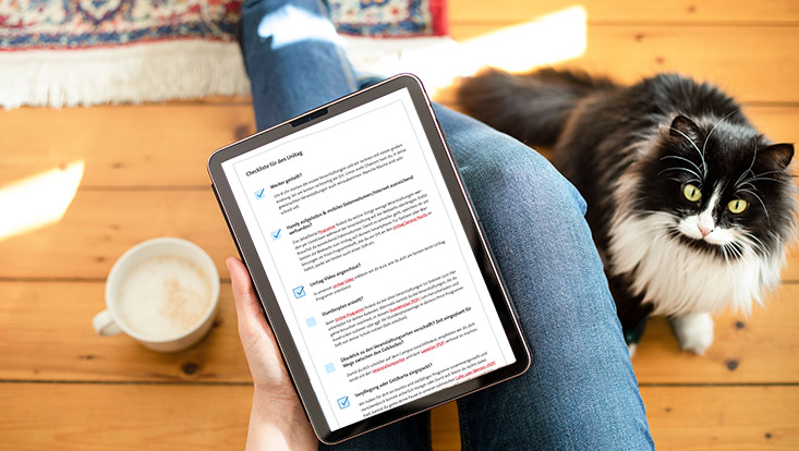 Ein Tablet mit einer Checkliste liegt auf den Knien, daneben sitzt eine Katze