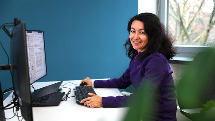 A woman with black hair sits at a computer and looks at the camera.