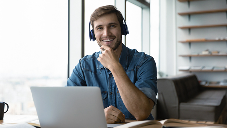 Portrait eines lächelnden Mannes mit Kopfhörern auf und sitzend am Schreibtisch vor einem Laptop