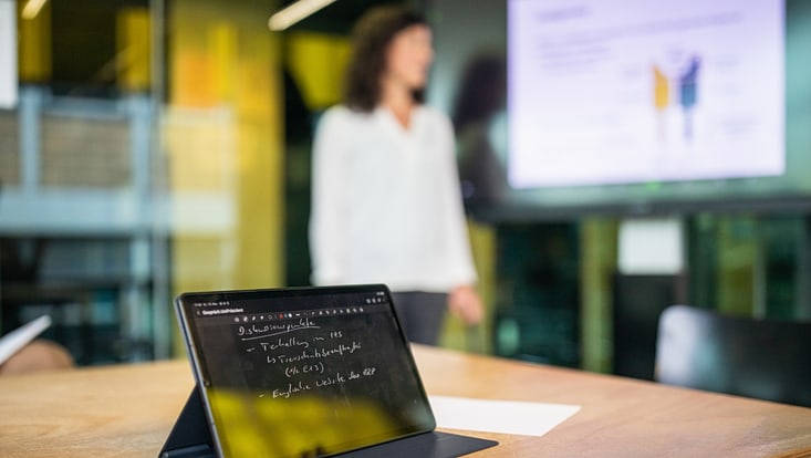 ein verschwommen dargestellte Person im Bildhintergrund hält eine Präsentation, im Vordergrund ist ein Tablet zu sehen