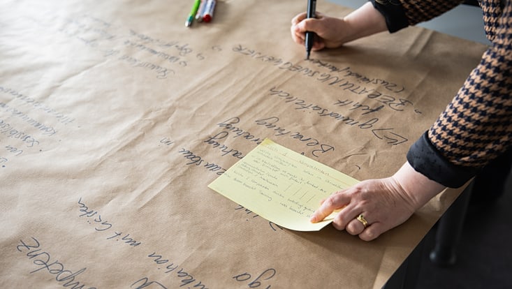 eine Person schreibt auf einem großen Stück Papier, hält eine Kartekarte in der anderen Hand (nur die Hände sind sichtbar)