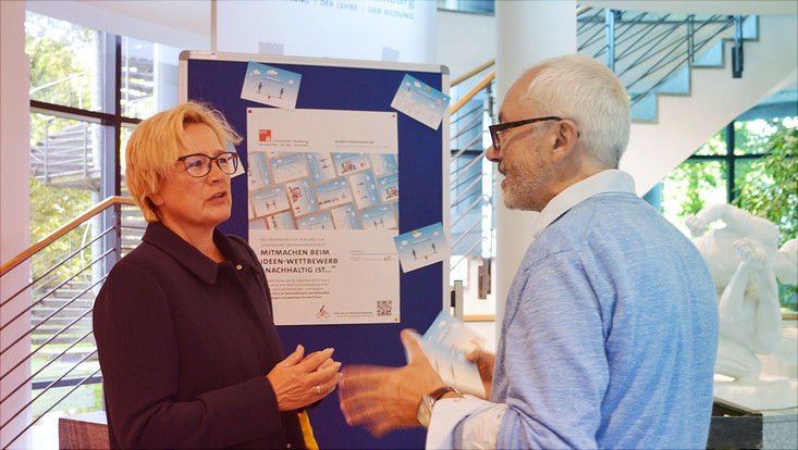 Zwei Menschen diskutieren im Foyer (Mittelweg). Im Hintergrund hängen ein Plakat und Postkarten zum Wettbwerb.