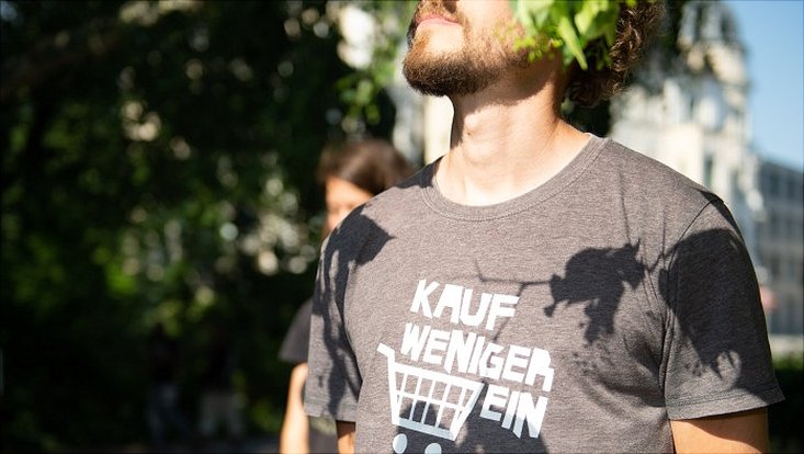 junger Mann mit rotem Vollbart schaut in den Himmel und trägt T-Shirt mit Aufschrift "Kauf weniger ein"