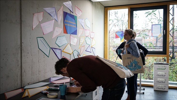 Ein Teilnehmer schreibt eine Karte für eine Feedbackwand, die von zwei Teilnehmerinnen betrachtet wird.