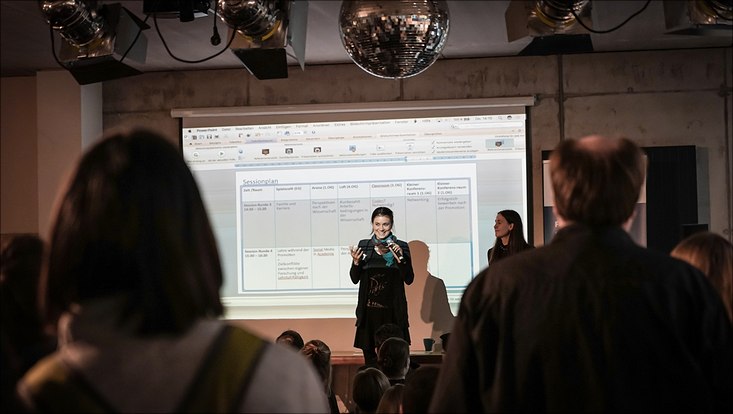 Auf der Bühne steht eine junge Frau mit einem Mikrofon in der Hand. Hinter ihr ist mithilfe eines Beamers der Sessionplan auf eine Leinwand geworfen.